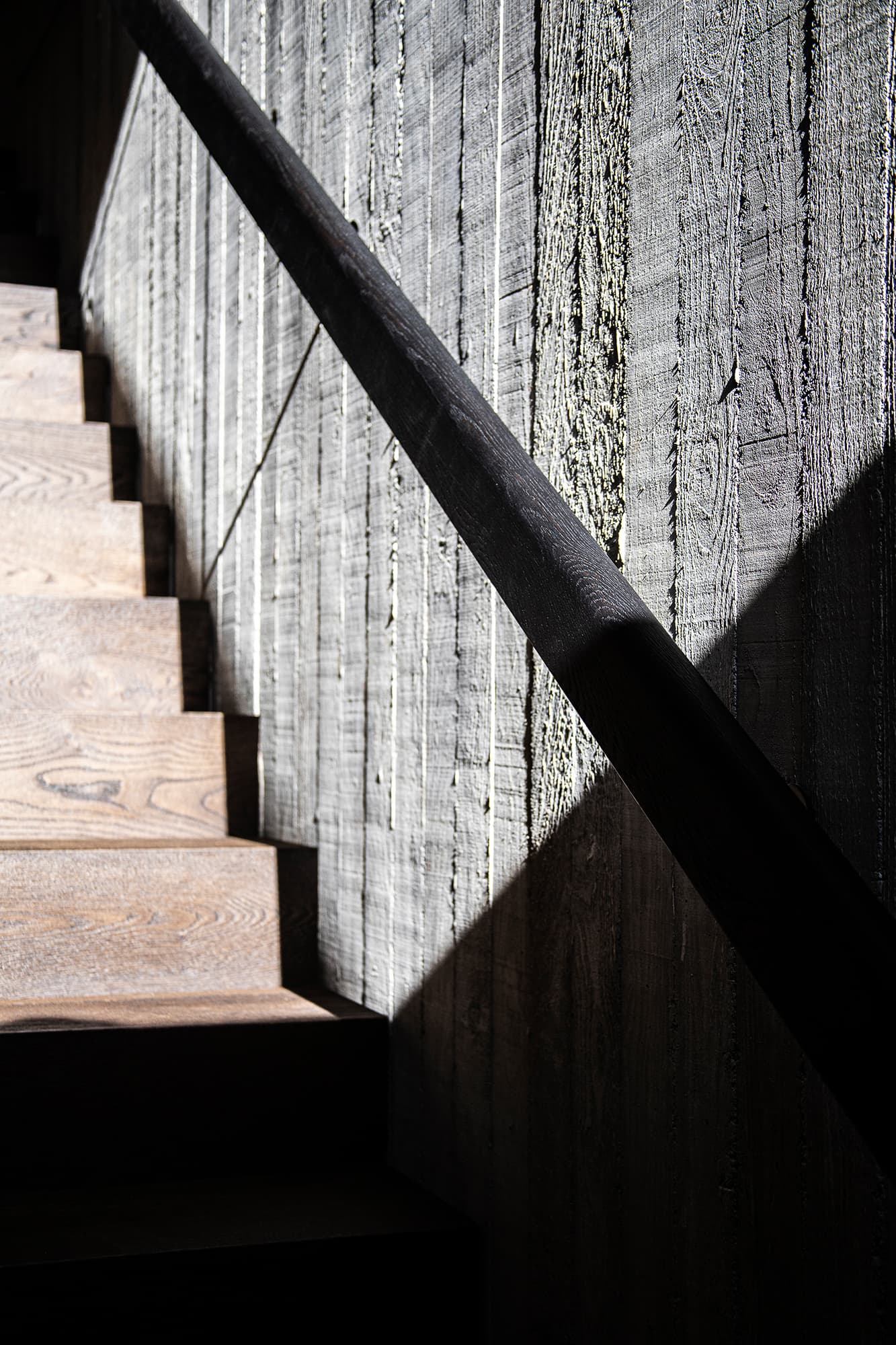 Alpenloge – Treppe mit dunklem Holzhandlauf vor strukturierter Holzwand im Streiflicht, Foto Michael Schott